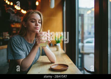 Giovane bionda in un caffè. Tiene una tazza di tè fragrante nelle sue mani e riposa Foto Stock