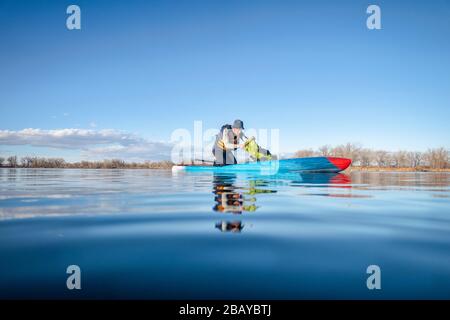 Il rematore maschile senior su una tavola da paddleboard sta guardando all'interno di una borsa a secco, un lago calmo in Colorado, scenario invernale o primaverile, azione ad angolo basso ca Foto Stock