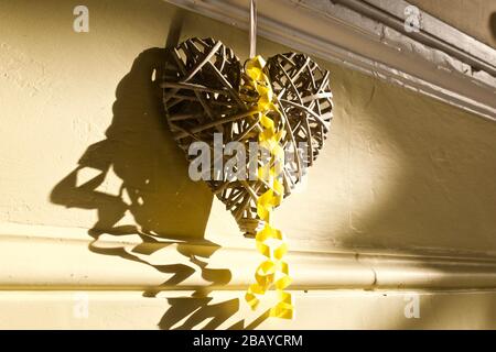 Cuore in vimini con nastro giallo, appeso a parete con luce e ombra interessanti Foto Stock