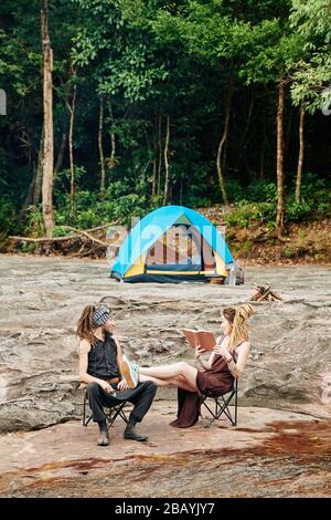 Donna che mette le gambe su giri di marito e di lettura lui un libro quando sono campeggio in foresta Foto Stock