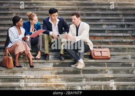 Gruppo di persone aziendali seri che discutono nuove tendenze e tendenze quando si siedono su gradini e guardano video su tablet digitali Foto Stock