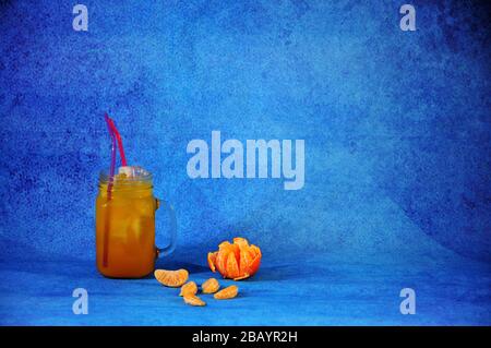 Una tazza di succo di mandarino con ghiaccio e cannucce accanto a pelati e tutta la frutta e fette di mandarino su uno sfondo blu. Close-up. Foto Stock