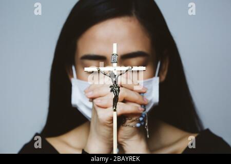 Donna che indossa maschera chirurgica che prega con rosario. Religione cristiana speranza concetto. Foto Stock