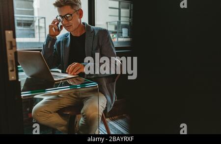 Uomo d'affari che parla con il cellulare e guarda il suo portatile. Uomo seduto alla sua scrivania di lavoro sul computer portatile e discutere sul telefono. Foto Stock