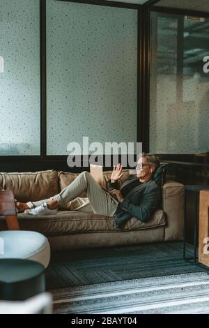 Uomo di mezza età in videochiamata utilizzando un computer portatile nella sala ufficio. Uomo d'affari sorridente sdraiato nella lounge dell'ufficio facendo gesti con una videochiamata. Foto Stock