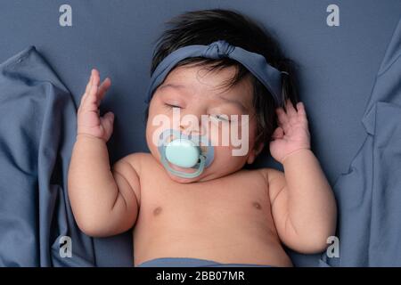 Bambino neonato che dorme a metà con capelli scuri con fasciatesta che si posa su uno sfondo blu con un succhietto in bocca. Foto Stock