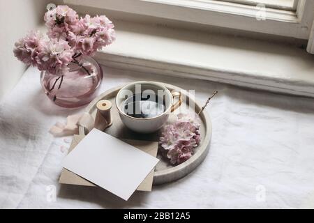 Primavera ancora vita scena. Biglietti d'auguri mockup, busta, vassoio in marmo, tazza di caffè, foto vintage in stile femminile. Composizione floreale con sak rosa Foto Stock