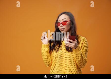 Donna asiatica in occhiali e con telefono in mano vape fumo su sfondo arancione vuoto in studio Foto Stock