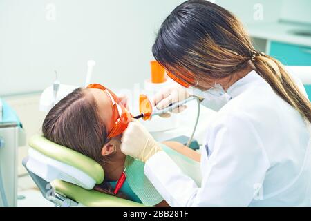 Ragazza bambino al medico. Il dentista posiziona un riempimento su un dente con la lampada di polimerizzazione dentale in cavità orale. Su sfondo clinico Foto Stock