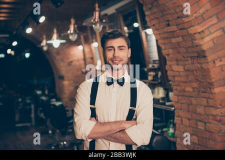 Attraente giovane bruna bearded guy in un barbiere, in piedi con le mani incrociate, sorridente, vestito di classe e stile retrò Foto Stock