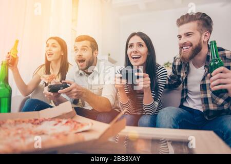 Gli amici eccitati stanno giocando ai videogiochi a casa seduti sul divano e facendo pizza con birra. Hanno un ottimo fine settimana in una bella compagnia al chiuso Foto Stock