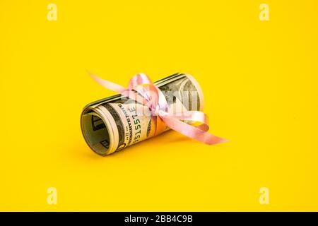 Piccolo regalo del dollaro, pacchetto delle fatture di cento dollari legati con un nastro rosa. Dollari isolati su sfondo giallo Foto Stock