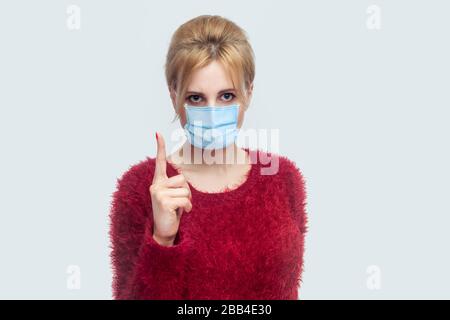 Ritratto di una giovane donna seria con maschera chirurgica medica in blusa rossa in piedi e guardando la fotocamera con un dito di avvertimento e allarmante sulla salute Foto Stock