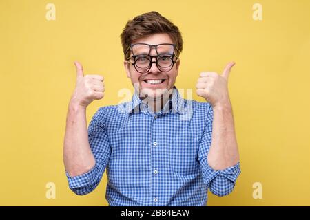 Un uomo in una camicia blu cerca sopra, seleziona gli occhiali per la visione che mostra il pollice in su che approva la scelta. Studio girato su yello muro. Foto Stock