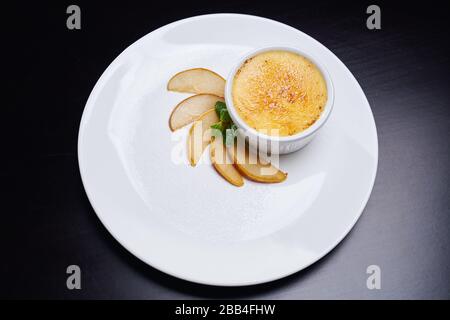 Vista dall'alto della piastra bianca con fette di pera fresca, menta, zucchero a velo e una piccola ciotola con cheesecake. Delizioso dessert al tavolo nel ristorante. Concetto di cucina. Foto Stock