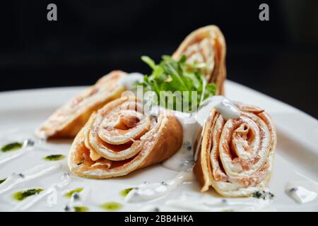 Chiudete quattro panini a fette con pasta e salmone serviti con rucola per insalata, pesto e panna acida su piatto bianco. Piatto saporito isolato su un tavolo di legno nero nel ristorante. Concetto di cucina. Foto Stock