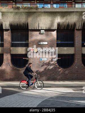Il Barbican, Silk Street, Londra. Un ciclista su una bici a noleggio con marchio Santander passa davanti all'architettura brutalista della tenuta Barbican. Foto Stock