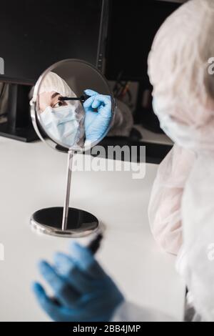 Ragazza in tuta trasparente bianca protettiva, guanti di gomma blu, maschera medica si siede a casa del tavolo e dipinge ciglia con mascara allo specchio durante Foto Stock