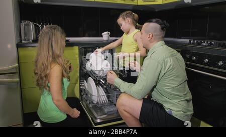 Madre e padre che insegnano a figlia imparare ad usare la lavastoviglie. Padrona ragazza bambini carico mettere piatti sporchi in lavastoviglie automatica. Foto Stock