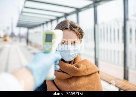 Misurazione della temperatura con termometro a infrarossi di una giovane donna in maschera facciale a un punto di controllo durante un'epidemia all'aperto. Concetto di prevenzione della diffusione del virus Foto Stock