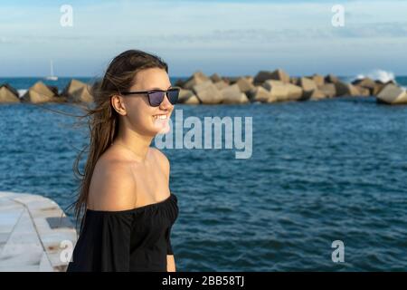 Una giovane bella donna felice con i capelli lunghi in piedi sul lungomare mentre guardando la macchina fotografica Foto Stock