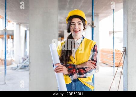 Il costruttore sicuro dell'ingegnere della ragazza si alza ad un luogo di costruzione che tiene la carta con i disegni Foto Stock