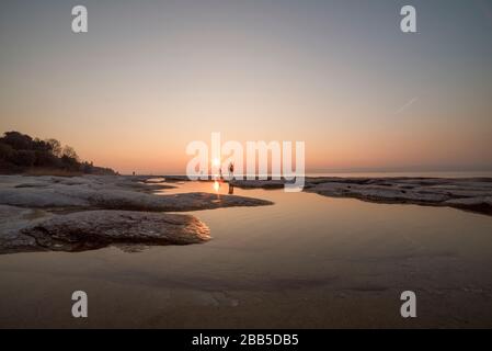 spiaggia di sirmione di notte e tramonto sul lago di Garda con rocce piatte sulla spiaggia e acqua e sole in l'orizzonte Foto Stock