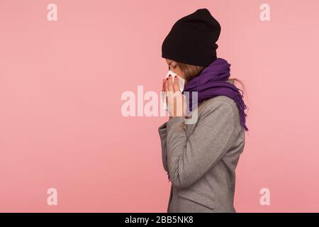 Influenza stagionale. Vista laterale della donna malata di influenza in caldo cappello e sciarpa che soffia naso nel tessuto, starnuti che soffrono sintomi allergici, sensazione di non sano. Foto Stock