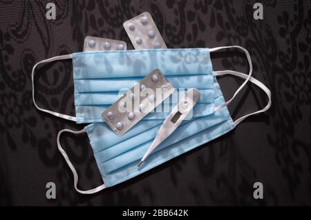Compresse, termometro e due maschere facciali monouso per uso medico blu sullo sfondo scuro con ornamenti ombreggiati contro il virus covid-2019. Cura di Heath Foto Stock