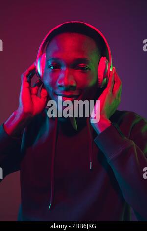 Ritratto uomo afroamericano con cuffie su sfondo gradiente studio in luce neon. Bel modello maschile cantando, ballando. Concetto di emozioni umane, espressione facciale, vendite, pubblicità, inclusione. Foto Stock