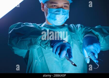 primo piano ritratto di un medico caucasico chirurgo, in un abito sterile, maschera, tenendo un bisturi e porta-ago con un ago, guidandoli all'opera Foto Stock