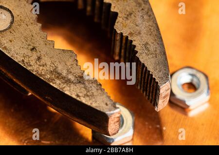 Una macro immagine delle ganasce di un paio di pinze arrugginite ben usate con diversi piccoli dadi in acciaio poggiati su una superficie di lavoro in rame usato ed ossidato. Caldo Foto Stock