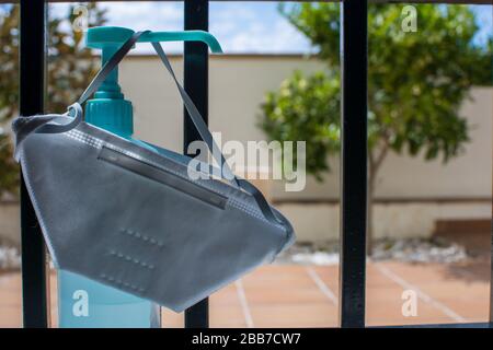 COVID-19 quarantena: Dispenser di gel igienizzante per le mani e maschera per la protezione aerea nella finestra di una casa con alberi e il cielo blu dietro le barre Foto Stock