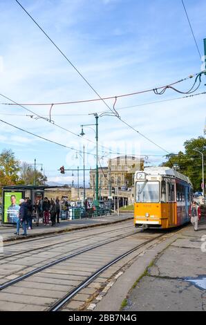 Budapest, Ungheria - 6 novembre 2019: Tram giallo alla stazione pubblica. Persone che camminano intorno. Il ponte della catena Szechenyi, l'edificio MTA e il centro storico sullo sfondo. Persone sulla strada. Giornata di sole. Foto Stock