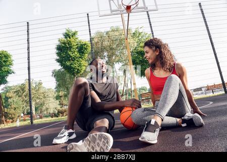 Attività all'aperto. Coppia africana seduta con palla sul campo di pallacanestro che parla sorridente allegro Foto Stock