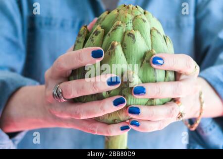 Donna con lucidalunghia blu brillante tiene un carciofo molto grande nelle sue mani, USA Foto Stock