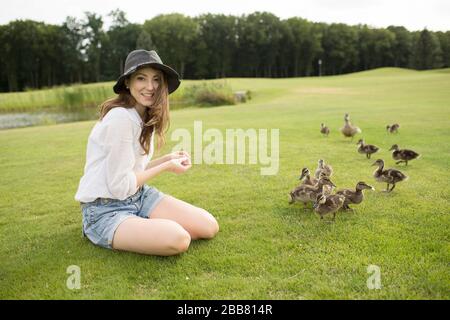 Bella giovane donna sedersi su erba verde oltre anatre piccole marrone. In mezzo al parco o al prato. Guarda la fotocamera e sorridi. Anatre di alimentazione Foto Stock