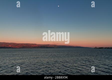 Veduta dello Stretto di Messina dal traghetto che lo attraversa, con una luna splenente e un faro sulla destra Foto Stock