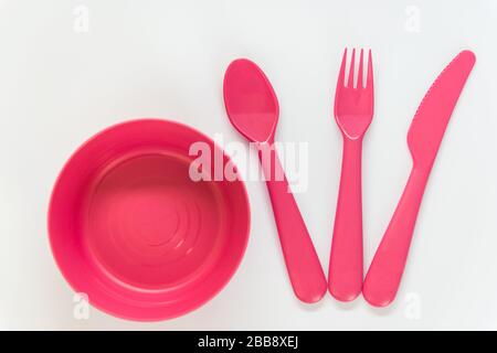 Cucchiaio di plastica rosa, coltello, forcella situata vicino alla piastra rosa sullo sfondo bianco. Disposizione piatta. Vista dall'alto. Foto Stock