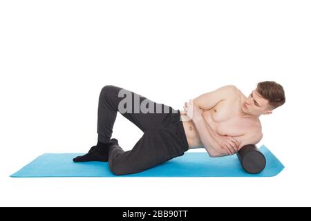 Il bell'uomo mostra esercizi, utilizzando un rullo in schiuma per un massaggio miofasciale sul tappetino da ginnastica in studio. Isolato su bianco. Foto Stock