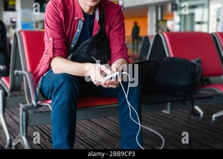 Man tourist hold smartphone ricarica via cavo usb da power bank portatile, seduto con bagagli al gate dell'aeroporto Foto Stock