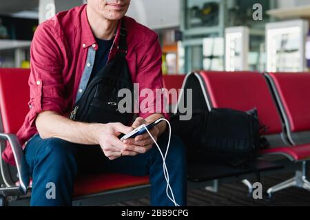 Man tourist hold smartphone ricarica via cavo usb da power bank portatile, seduto con bagagli al gate dell'aeroporto Foto Stock