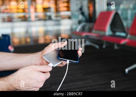 Il viaggiatore maschio ricarica il telefono cellulare con dispositivo di ricarica portatile tramite usb a sfondo sala aeroporto. Primo piano. Foto Stock