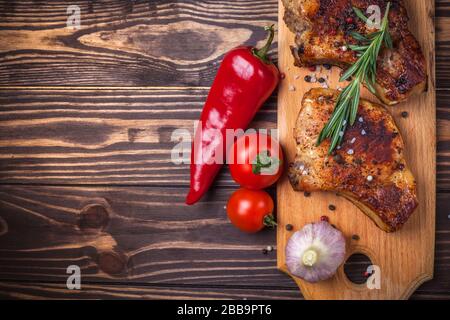 Bistecche di maiale alla griglia e verdure su un rustico tavolo di legno. Carne alla griglia sull'osso con spezie, aglio, peperoni, pomodori e rosmarino. Menu del ristorante. Vista dall'alto Foto Stock