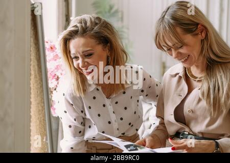 Due ragazze ridono discutendo le idee. Stilisti stilisti stilisti di stilisti che lavorano su un nuovo progetto in studio, utilizzando scrivania, riviste, schizzi Foto Stock