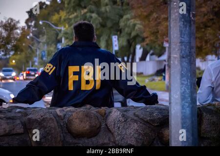 Agente FBI maschile che indossa un cappotto blu scuro con logo FBI che guarda lungo la strada con le auto al tramonto visto da dietro Foto Stock