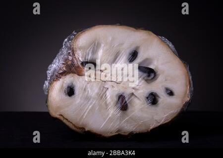 Avvolgimento in plastica riflettente intorno a un singolo Soursop o Graviola frutta aperta con semi neri in polpa bianca. Un'immagine in studio a basso contenuto chiave di cibi freschi confezionati. Foto Stock