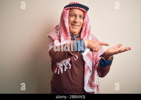 Anziano bell'uomo arabo che indossa il keffiyeh in piedi su sfondo bianco isolato stupito e sorridente alla macchina fotografica mentre presenta con mano e poin Foto Stock