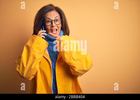 Donna in età media che indossa un impermeabile giallo che chiama utilizzando lo smartphone, è molto felice di puntare con mano e dito Foto Stock