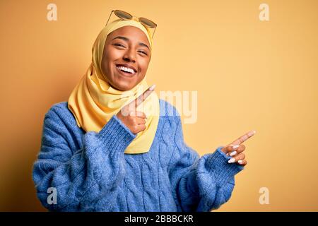 Giovane studentessa afroamericana indossando hijab musulmano giallo e occhiali da sole sorridenti e guardando la macchina fotografica che punta con due mani e dita Foto Stock